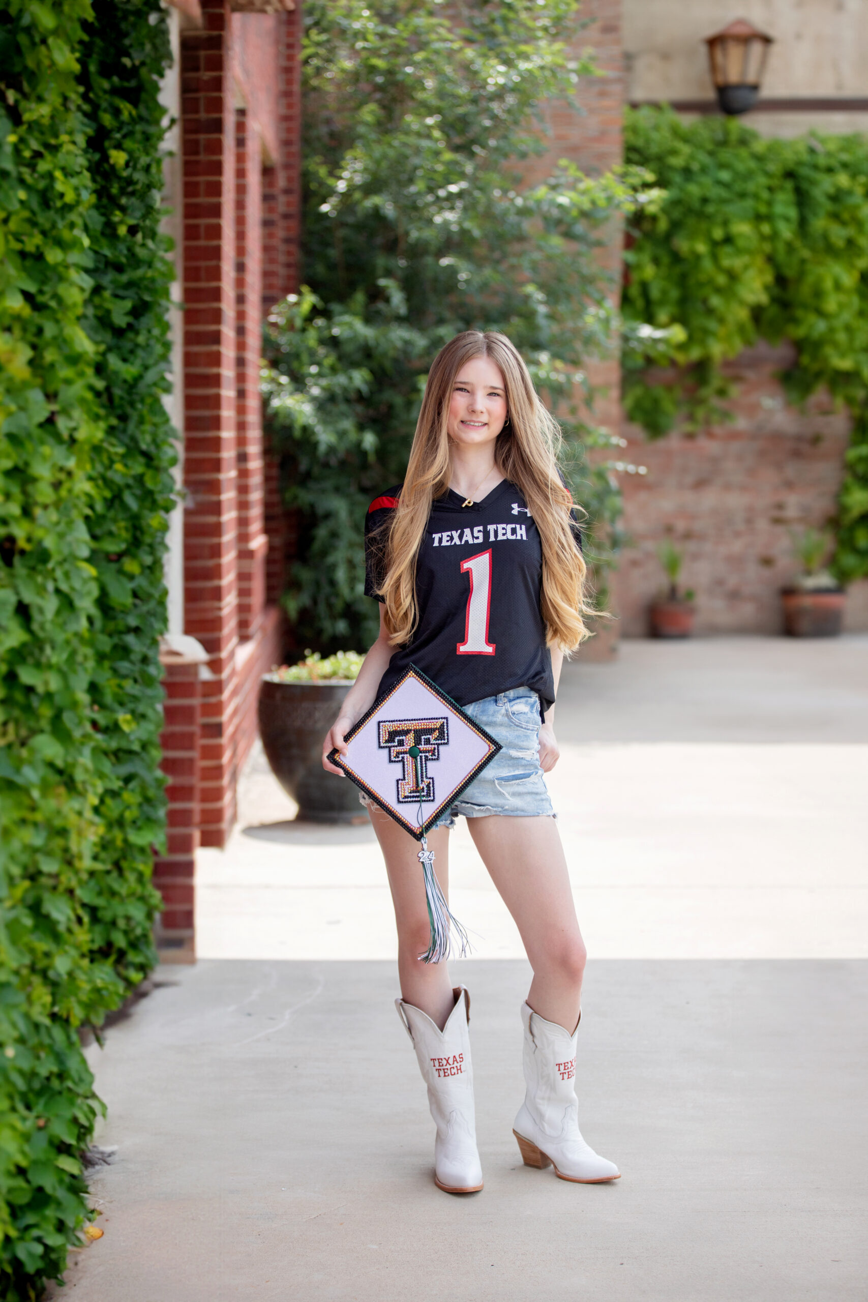 Graduate in a Texas Tech jersey holding her decorated cap in a brick courtyard
